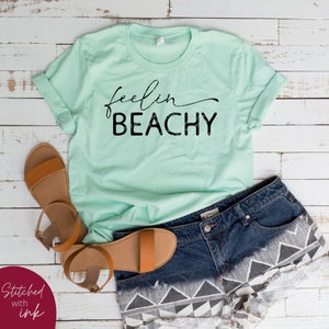 May include: A light green t-shirt with the text "feelin BEACHY" in black lettering. The shirt is laid out on a white wooden surface with a pair of brown sandals and denim shorts with a black and white geometric pattern.