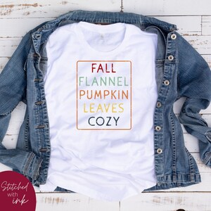 Puede incluir: Camiseta blanca con un gr&aacute;fico de estilo vintage que dice "Fall Flannel Pumpkin Leaves Cozy" en un rect&aacute;ngulo con un borde desgastado.