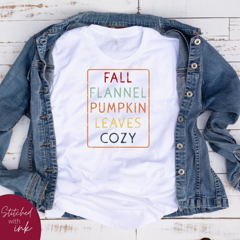 Puede incluir: Camiseta blanca con un gr&aacute;fico de estilo vintage que dice "Fall Flannel Pumpkin Leaves Cozy" en un rect&aacute;ngulo con un borde desgastado.