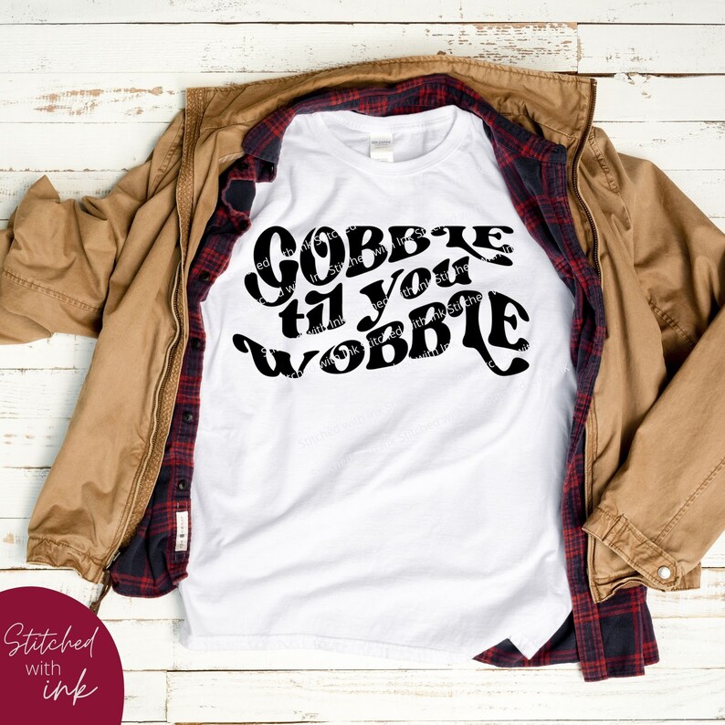 May include: White t-shirt with black text that reads "Gobble til you wobble". The shirt is laid on a wooden surface with a brown jacket and a red and black plaid shirt on top of it.