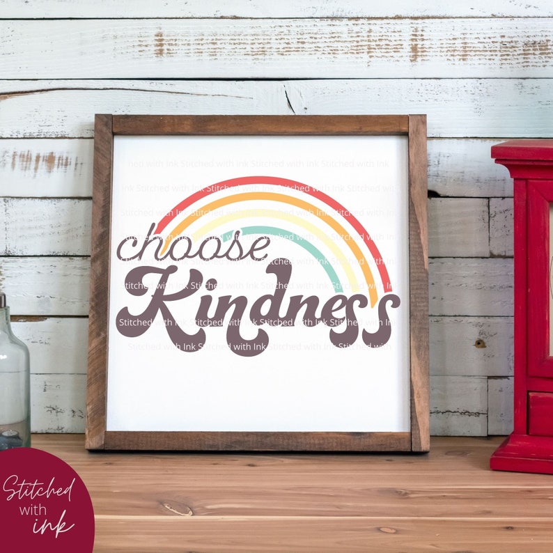 May include: A framed wall art piece with a white background and a rainbow design. The words "choose kindness" are written in a stylized font. The frame is made of brown wood. The artwork is displayed on a wooden surface.