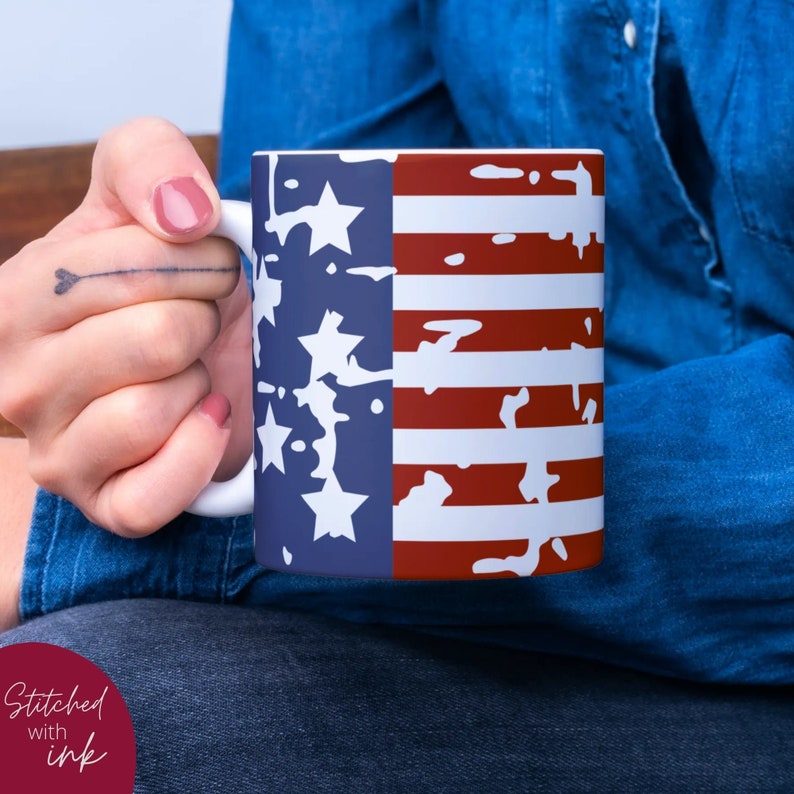 K&ouml;nnte beinhalten: Eine wei&szlig;e Keramiktasse mit einem abgenutzten Design der amerikanischen Flagge. Die Flagge ist blau, wei&szlig; und rot mit wei&szlig;en Sternen auf dem blauen Teil. Die Tasse wird von einer Person gehalten, die ein blaues Jeanshemd tr&auml;gt.