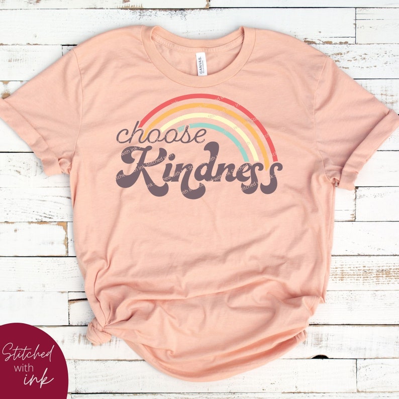 May include: A peach-colored t-shirt with a rainbow graphic and the words "choose Kindness" in a retro font. The shirt is made of soft material and is suitable for casual wear.