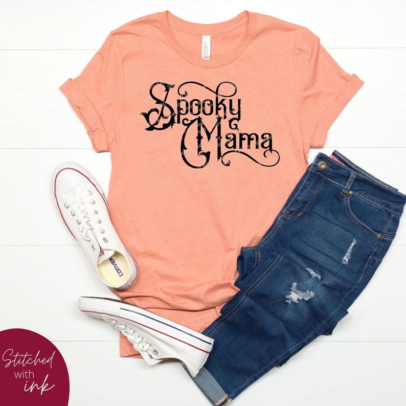 May include: A peach colored t-shirt with the text "Spooky Mama" in black lettering. The shirt is styled with blue jeans and white sneakers.