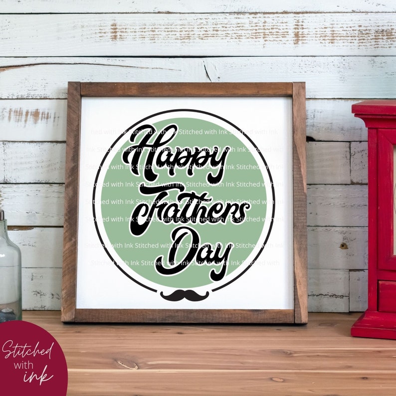 May include: A green and black sign with the text "Happy Fathers Day" in a circle with a black mustache underneath. The sign is framed in a brown wood frame.