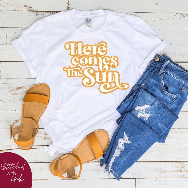 May include: White t-shirt with the text "Here comes the sun" in yellow, styled with blue jeans and brown sandals.