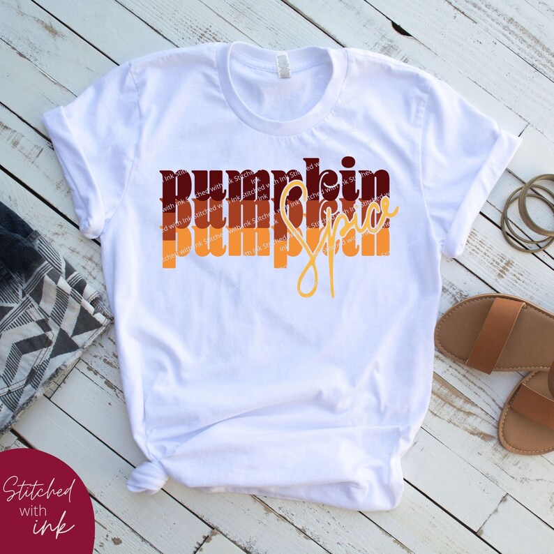 May include: White t-shirt with a graphic design that says "Pumpkin Spice" in a layered, retro style with orange, brown, and red colors.