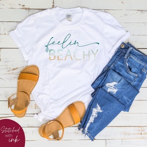 May include: White t-shirt with the text "feelin beachy" in a green, blue, and orange gradient. The shirt is laid out on a white wooden surface with a pair of brown sandals and blue jeans.