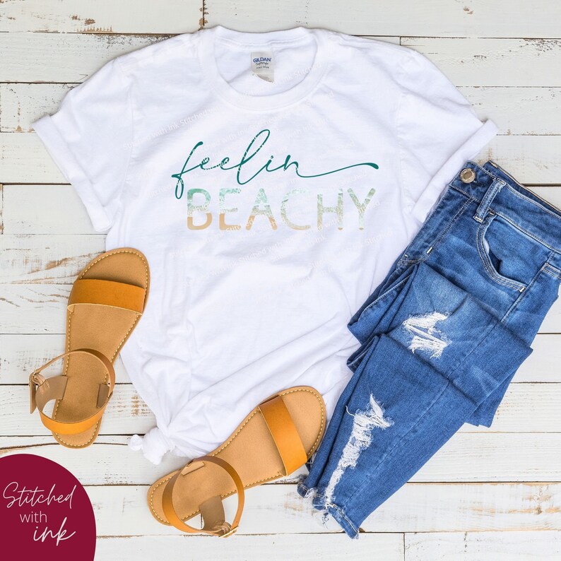 May include: White t-shirt with the text "feelin beachy" in a green, blue, and orange gradient. The shirt is laid out on a white wooden surface with a pair of brown sandals and blue jeans.
