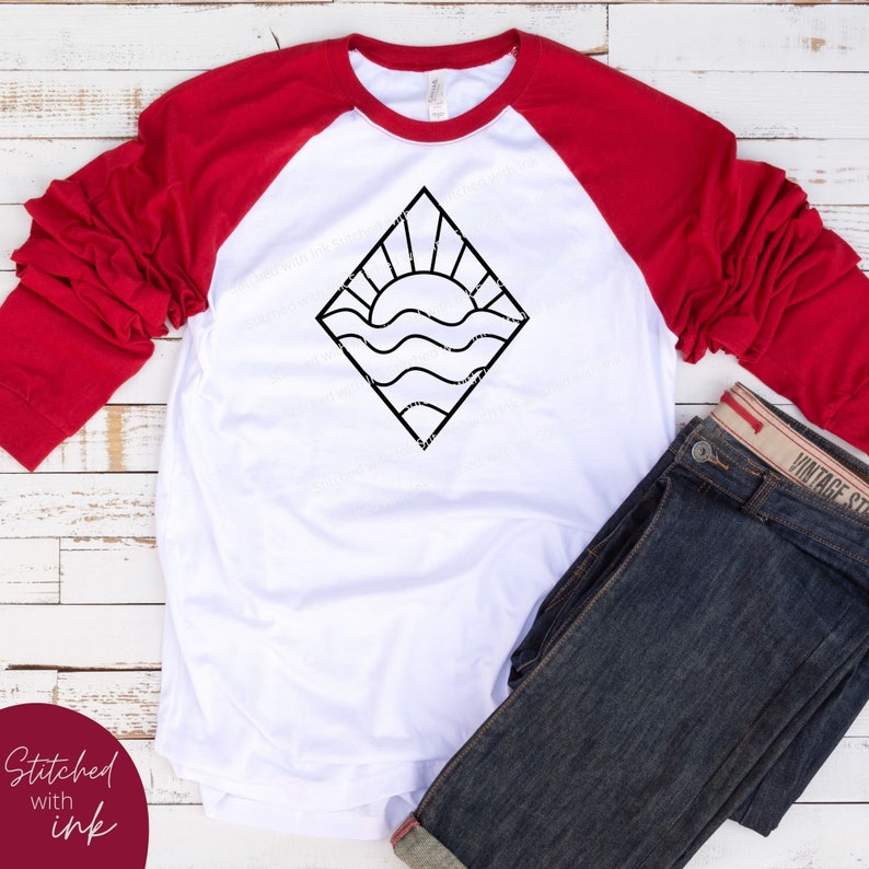 May include: A white and red baseball tee shirt with a black graphic of a sun setting over waves inside a diamond shape. The shirt is laying on a white wooden surface with a pair of blue jeans.