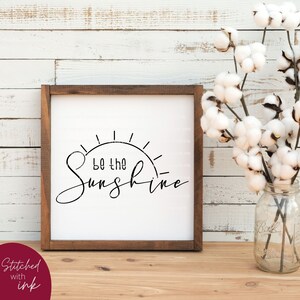 Pu&ograve; includere: Un cartello incorniciato con sfondo bianco e testo nero che recita "Be the Sunshine" con una grafica solare stilizzata. Il cartello &egrave; in una cornice di legno e si trova su un tavolo di legno.