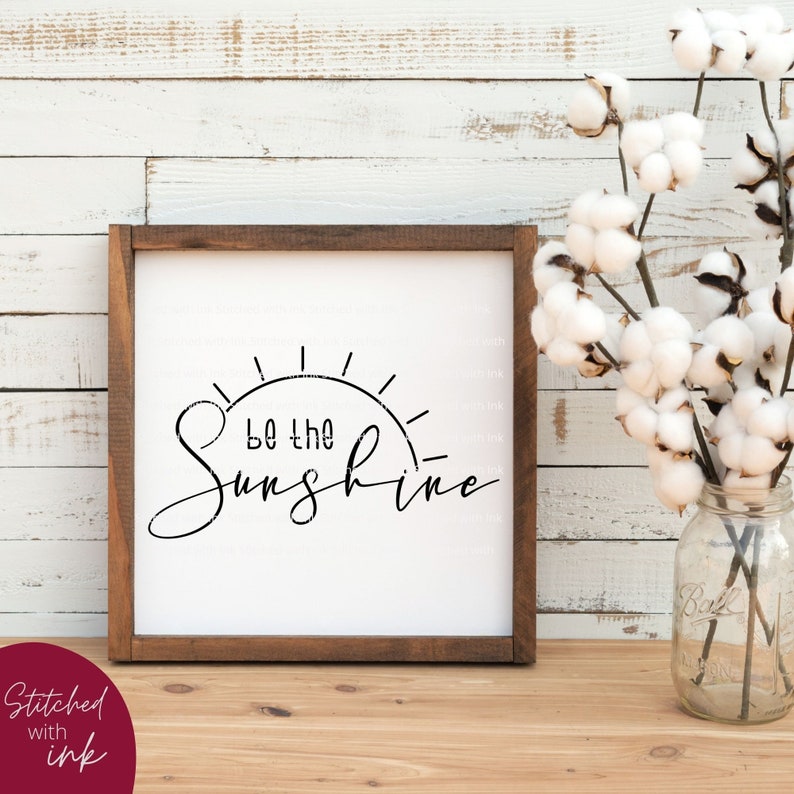 Pu&ograve; includere: Un cartello incorniciato con sfondo bianco e testo nero che recita "Be the Sunshine" con una grafica solare stilizzata. Il cartello &egrave; in una cornice di legno e si trova su un tavolo di legno.