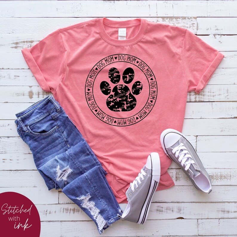 Op de afbeelding: Een roze T-shirt met een zwart pootprintontwerp in een cirkel. De cirkel heeft de tekst "Dog Mom" eromheen herhaald. Het shirt ligt op een wit houten oppervlak met een blauwe spijkerbroek en grijze sneakers.