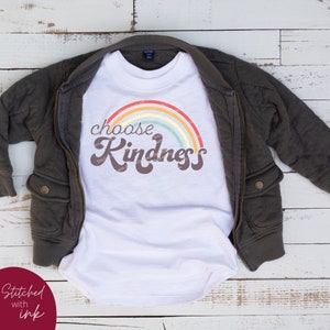 May include: A white t-shirt with a rainbow graphic and the words "choose Kindness" in brown script. The shirt is displayed on a dark gray jacket with a zipper and pockets. The words "Stitched with ink" are in the lower left corner.