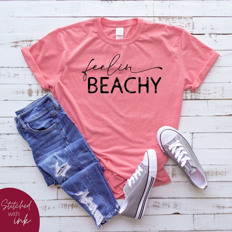 May include: A pink t-shirt with the text "feelin BEACHY" in black lettering. The shirt is laid out on a white wooden surface with a pair of blue jeans and gray sneakers.