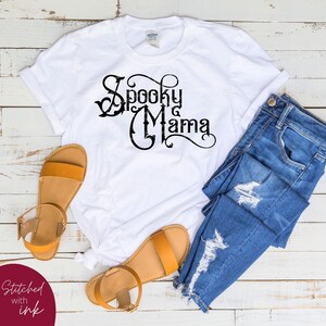 May include: White t-shirt with black text that reads "Spooky Mama" in a decorative font. The shirt is laid flat on a white wooden surface with a pair of brown sandals and blue jeans.