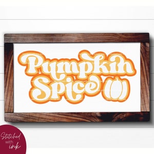 May include: A white sign with a brown wooden frame, featuring the text "Pumpkin Spice" in a retro orange and yellow font. The text is layered with a white outline and a shadow effect.