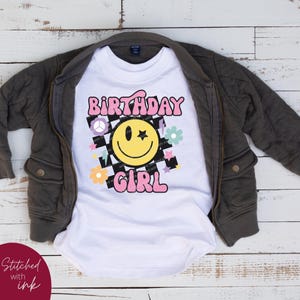 Op de afbeelding: Wit T-shirt met een roze "Birthday Girl"-grafiek met een gele smiley met een ster als oog, een vredesteken en bloemen op een zwart-wit geruite achtergrond.