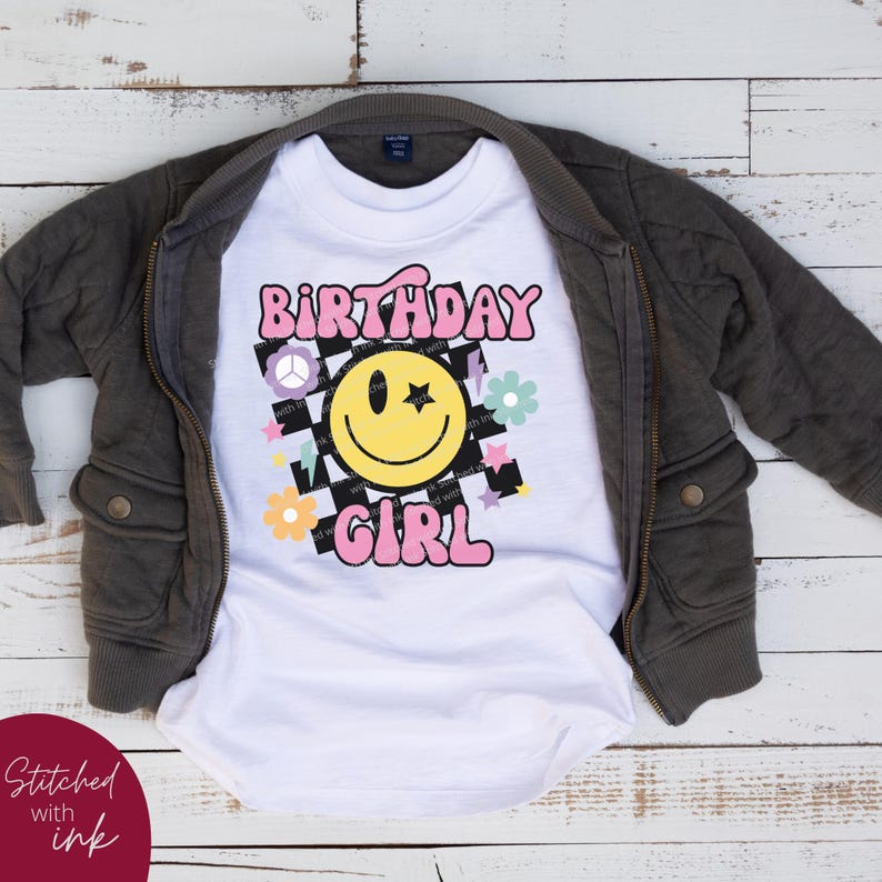Op de afbeelding: Wit T-shirt met een roze "Birthday Girl"-grafiek met een gele smiley met een ster als oog, een vredesteken en bloemen op een zwart-wit geruite achtergrond.