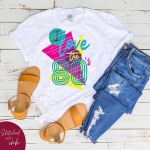 May include: A white t-shirt featuring a graphic design that reads "I love the 80's" in a colourful retro style. The design incorporates a pink, yellow and blue geometric pattern.