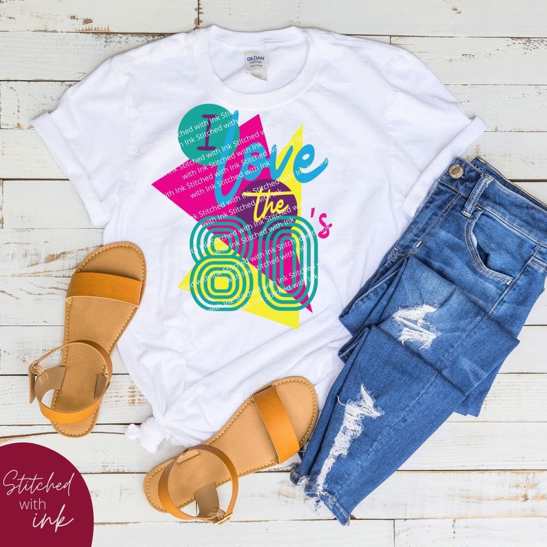 May include: A white t-shirt featuring a graphic design that reads "I love the 80's" in a colourful retro style. The design incorporates a pink, yellow and blue geometric pattern.