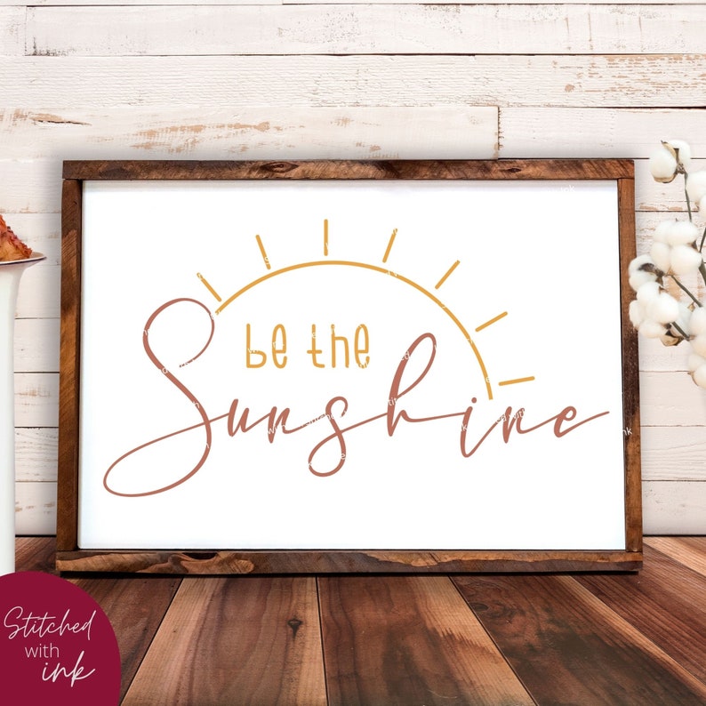 Pu&ograve; includere: Un cartello incorniciato di bianco con il testo "Be the Sunshine" in lettere marroni e dorate. Le lettere sono in un font corsivo. Il cartello &egrave; su una superficie di legno.