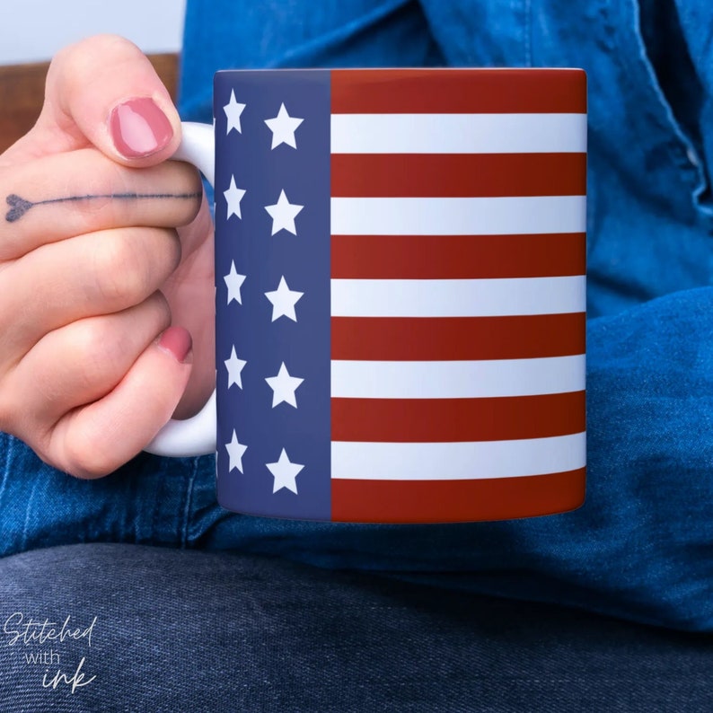 K&ouml;nnte beinhalten: Eine wei&szlig;e Keramiktasse mit einem rot-wei&szlig;-blauen amerikanischen Flaggenmuster. Die Tasse wird von einer Hand gehalten.