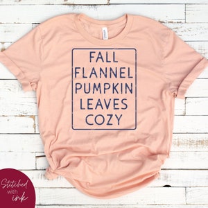 Puede incluir: Una camiseta de color melocot&oacute;n con un gr&aacute;fico azul desgastado que dice "Fall Flannel Pumpkin Leaves Cozy".