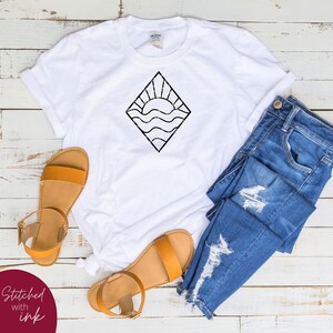 May include: White t-shirt with a black graphic of a sun setting over waves inside a diamond shape. The t-shirt is laid out on a wooden surface with a pair of brown sandals and blue jeans.