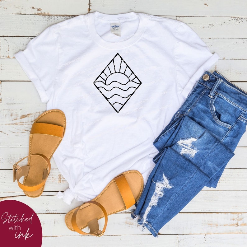 May include: White t-shirt with a black graphic of a sun setting over waves inside a diamond shape. The t-shirt is laid out on a wooden surface with a pair of brown sandals and blue jeans.
