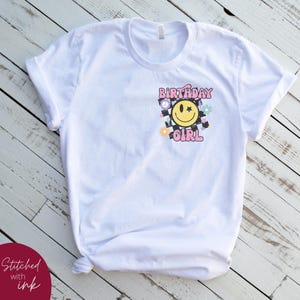 Op de afbeelding: Wit T-shirt met een roze en geel grafisch ontwerp met de tekst "Birthday Girl" met een smiley en een ster.