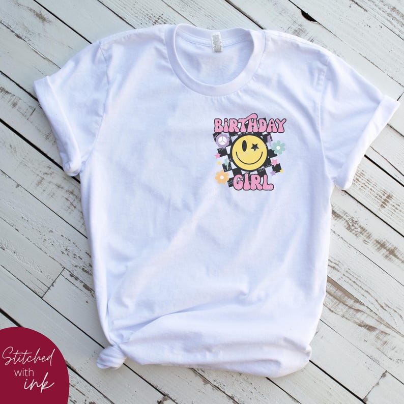 Op de afbeelding: Wit T-shirt met een roze en geel grafisch ontwerp met de tekst "Birthday Girl" met een smiley en een ster.