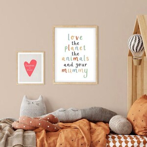Może przedstawiać: Pokój dziecięcy z drewnianym łóżkiem, beżowym kocem i dwoma obrazami w ramkach na ścianie. Obraz po lewej stronie głosi "Naucz się kochać", a obraz po prawej stronie głosi "Kochaj planetę, zwierzęta i mamę".