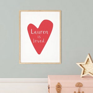 Może przedstawiać: Wydruk w ramie z czerwonym sercem i tekstem "Lauren is loved." Wydruk jest na białym tle i ma drewnianą ramę. Wydruk wisi na jasnoszarej ścianie.