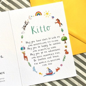 May include: A white card with a green circle around the name "Kitto" and a colourful illustration of a rainbow, sun, stars, animals, and a boat. The card has a message wishing the person a happy life with stars to wish on, mountains to climb, and adventures to go on.