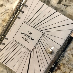 May include: A clear plastic binder with a white page inside. The page has a black and white design with numbers 1 through 31 and the text "I'm Grateful For..."  The binder has a silver ring mechanism and a silver button.