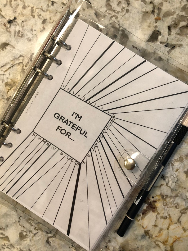 May include: A clear plastic binder with a white page inside. The page has a black and white design with numbers 1 through 31 and the text "I'm Grateful For..."  The binder has a silver ring mechanism and a silver button.