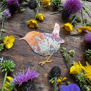 Könnte beinhalten: Metall-Vogel-Silhouette mit verwitterter, mehrfarbiger Oberfläche, umgeben von Wildblumen in Gelb-, Lila- und Grüntönen. Der Vogel hat eine rostig-orange und blau-grüne Patina, vor einem rustikalen Holzgrund.