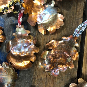 May include: A collection of copper oak leaf ornaments with a rustic, aged finish. Each leaf is unique and has a different patina. The leaves are strung together with fabric ribbon.