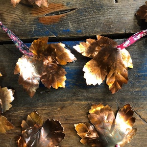 May include: A collection of copper leaf ornaments with a floral pattern. The leaves are arranged on a wooden surface and are connected by a red and white ribbon.