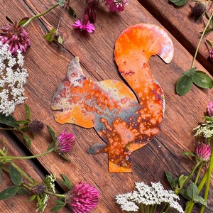 Könnte beinhalten: Metall-Eichhörnchen-Silhouette mit orangefarbenem, braunem und blauem, gesprenkeltem Finish. Das Eichhörnchen ist von rosa und weißen Wildblumen auf einer rustikalen Holzoberfläche umgeben. Ein dekorativer Artikel.