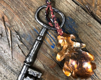 May include: An antique silver key with a copper leaf charm attached to a red and black fabric loop.