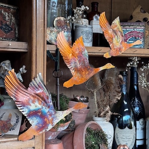 May include: Three metal goose sculptures in flight, with orange, red, and blue tones. The wings are outstretched, and the bodies are streamlined. The geese are displayed on a wooden shelf with other decorative items, including bottles and potted plants.