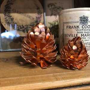 May include: Two copper pinecone-shaped candle holders. The larger holder is in the foreground and the smaller holder is in the background. Both holders have a shiny, metallic finish.