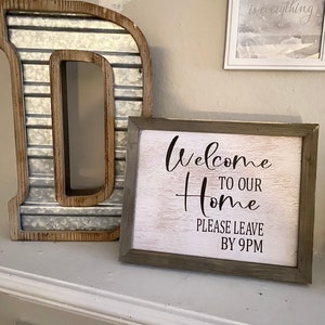 May include: A white wooden sign with a gray frame that says "Welcome to our Home Please Leave by 9PM" in black script font.