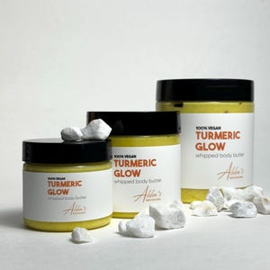 May include: Three jars of turmeric glow whipped body butter, labeled "100% Vegan Turmeric Glow Whipped Body Butter" with the brand name "Alba's" on the label. The jars are on a white surface with white rocks.