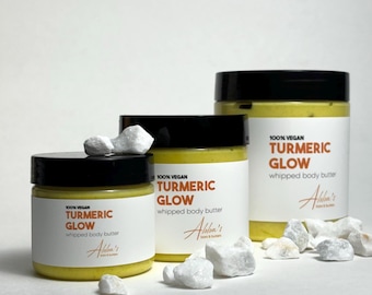 May include: Three jars of turmeric glow whipped body butter, labeled "100% Vegan Turmeric Glow Whipped Body Butter" with the brand name "Alba's" on the label. The jars are on a white surface with white rocks.