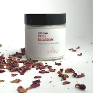 May include: A white jar with a black lid containing whipped body butter. The jar has a label that reads "100% Vegan Rose Blossom whipped body butter" and "Alba's Lotions & Butters". The jar is surrounded by dried rose petals on a white surface.