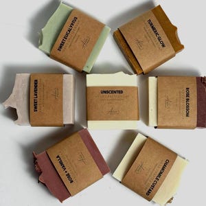 May include: Assortment of ten handmade soap bars with various scents, including Sweet Lavender, Sweet Eucalyptus, Turmeric Glow, Rose Blossom, Chamomile Custard, Rose Vanilla, and Unscented. Each bar is wrapped in brown paper with a label indicating the scent.