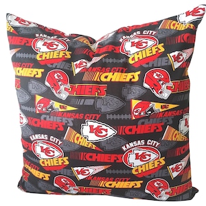 May include: A square pillow with a black background featuring a repeating pattern of red and yellow Kansas City Chiefs logos, helmets, and text. The pillow is filled and ready for use.